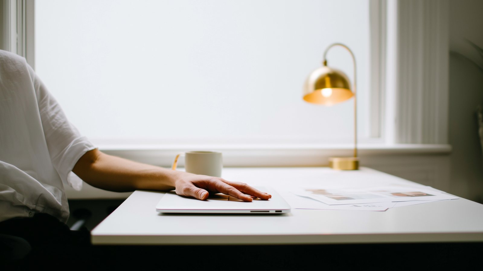 A hand resting on a closed laptop at a white desk, with printed pages and a coffee cup beside it