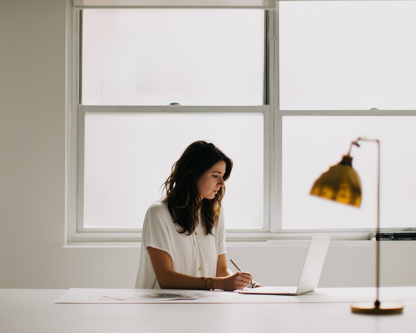 Someone writing at a large desk by a window, notebook open beside a laptop