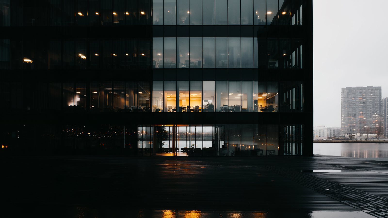 Een donker kantoorgebouw 's nachts aan het water, amberkleurig licht weerspiegeld op de kade